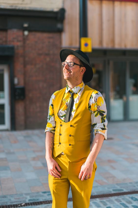 a colorful groom's look with a bold floral print shirt, a bold yellow waistcoat and pants, a black tie and a black hat
