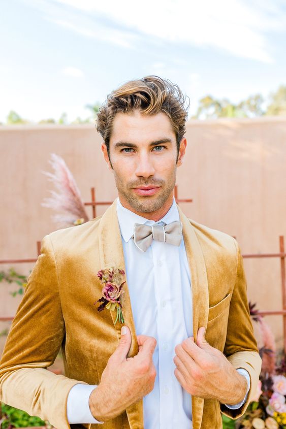 a glam groom's look with a white shirt, a grey bow tie, a mustard velvet blazer and a pink boutonniere for a glam wedding