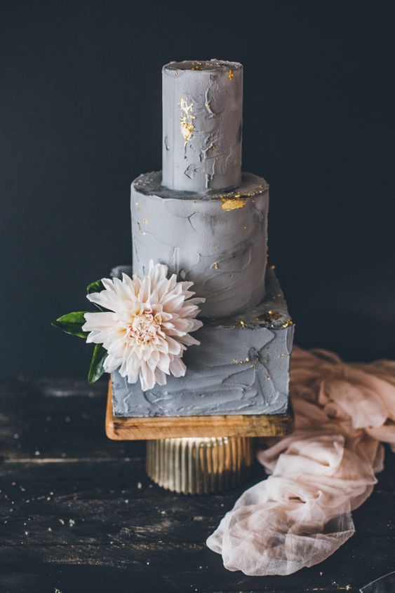 a grey textural wedding cake with tiers of different shapes and gold foil, a blush bloom with greenery for a modern wedding
