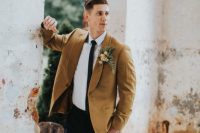 a modern fall groom’s look with a white shirt, a black tie and trousers, a mustard blazer and a bold boutonniere