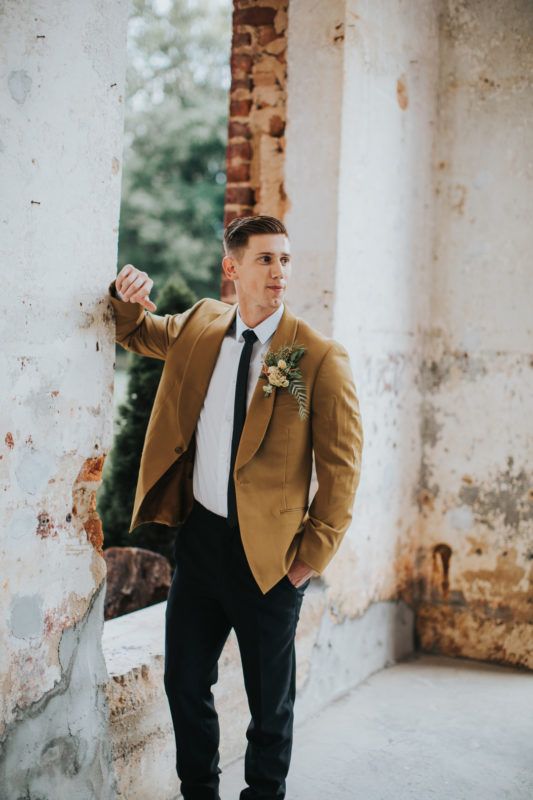 a modern fall groom's look with a white shirt, a black tie and trousers, a mustard blazer and a bold boutonniere