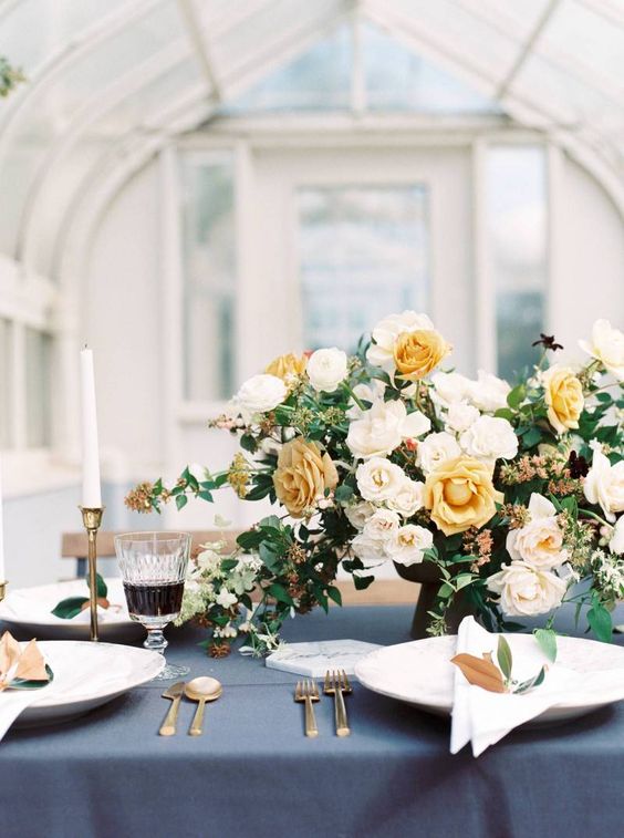 a refined wedding table setting with a slate grey tablecloth, white porcelain and napkins, white and yellow blooms and candles