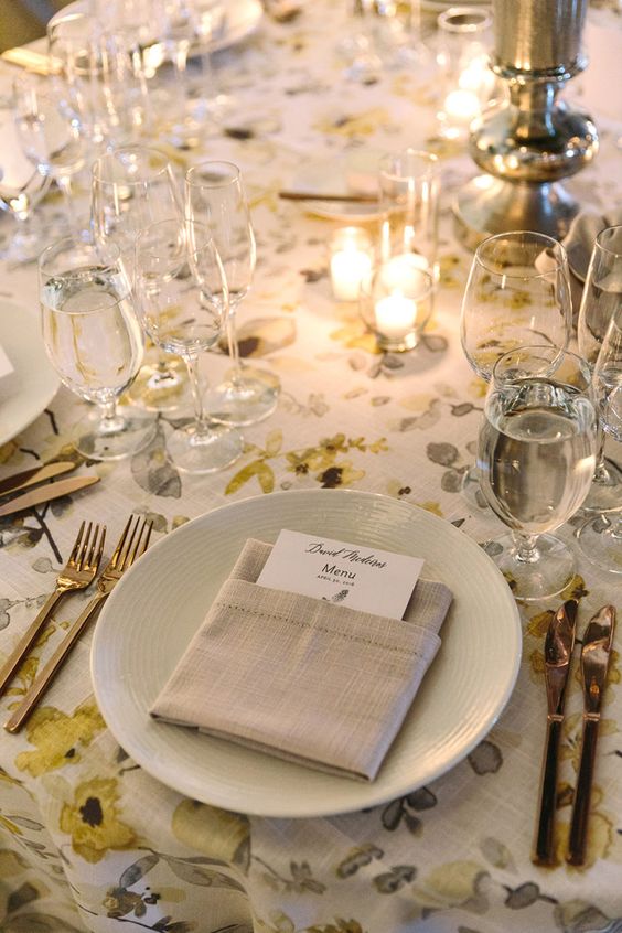 an elegant wedding tablescape with a floral tablecloth, neutral plates, grey napkins, glasses, candles and metallic cutlery