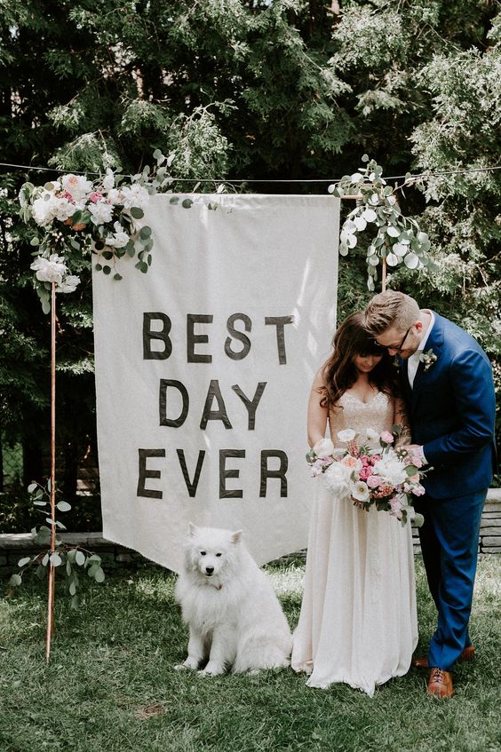 a backyard wedding arch done with blooms and greenery and a large banner and the couple's dog taking part in the wedding