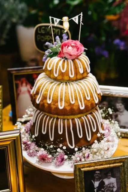 a bundt wedding cake with creamy drip, lilac and pink blooms, baby’s breath and a small banner topper for a summer wedding