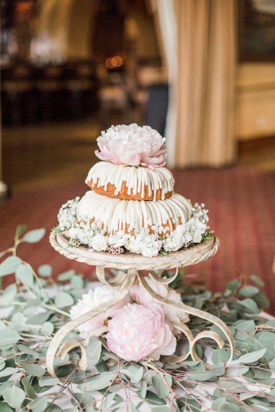 a cute two tier bundt wedding cake with creamy drip, a large blush bloom on top and white blooms that surround it