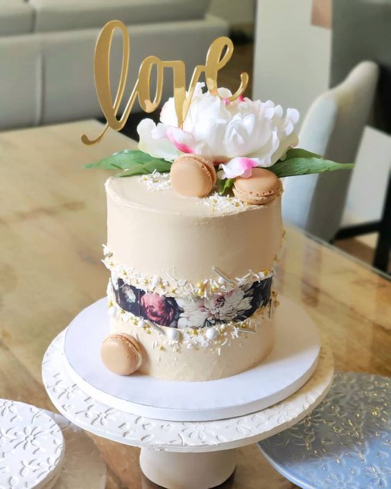 a neutral wedding cake with a dark floral fault line and some confetti and beads, topped with a single bloom and some macarons