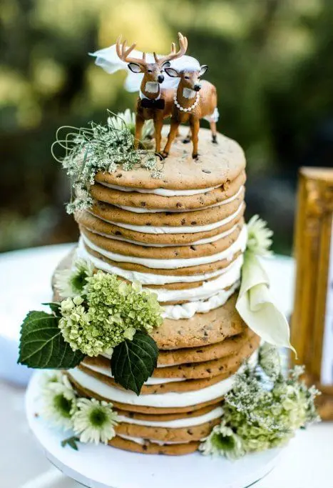 a chocolate chip cookie wedding cake with white filling and greenery decor plus fun deer toppers for a woodland wedding