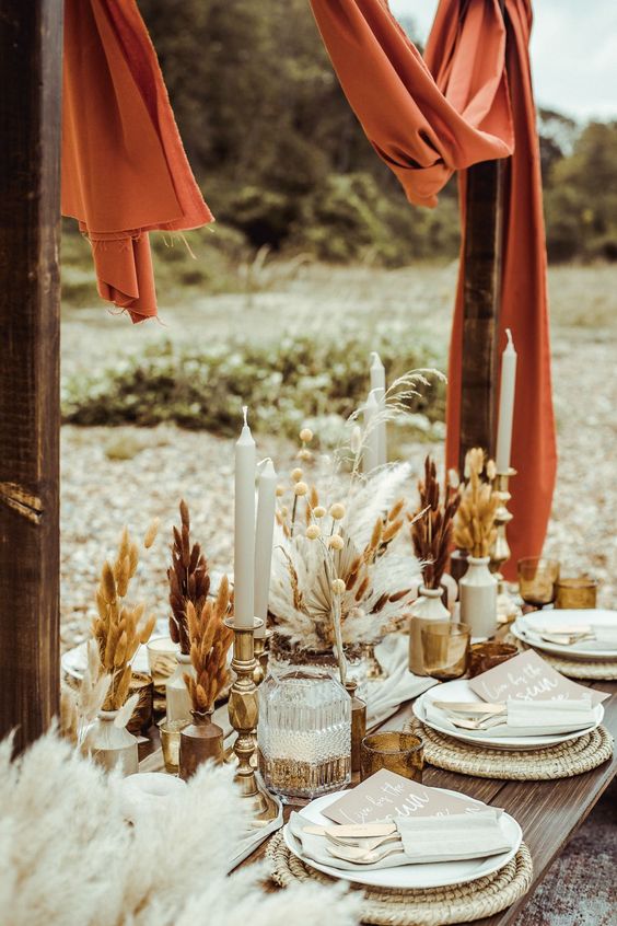 a beautiful boho wedding tablescape with woven placemats, grasses in vases, candles and rust fabric over the table