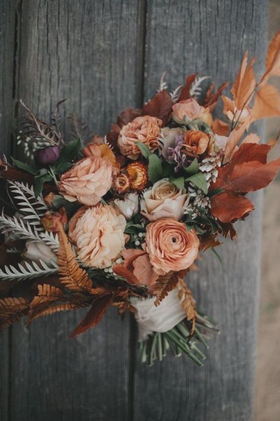 a beautiful fall wedding bouquet with rust fall leaves, greenery, pale greenery and peachy and orange blooms