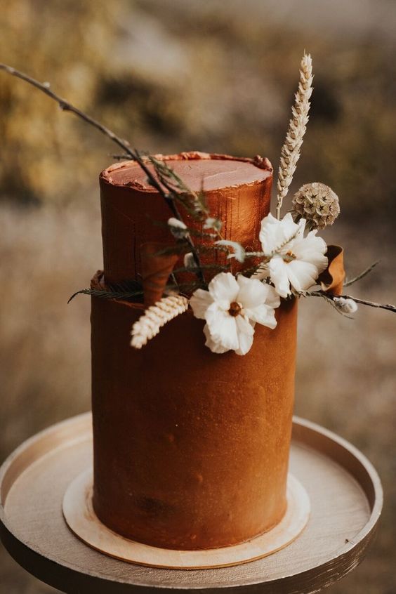 a rust-colored textural wedding cake with white blooms, grasses, greenery and a rough edge is a bold idea for a boho wedding