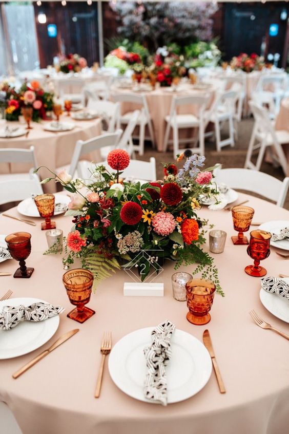 a beautiful colorful wedding centerpiece of greenery, burgundy, deep red, yellow and orange blooms, candles around and a clear acrylic table number