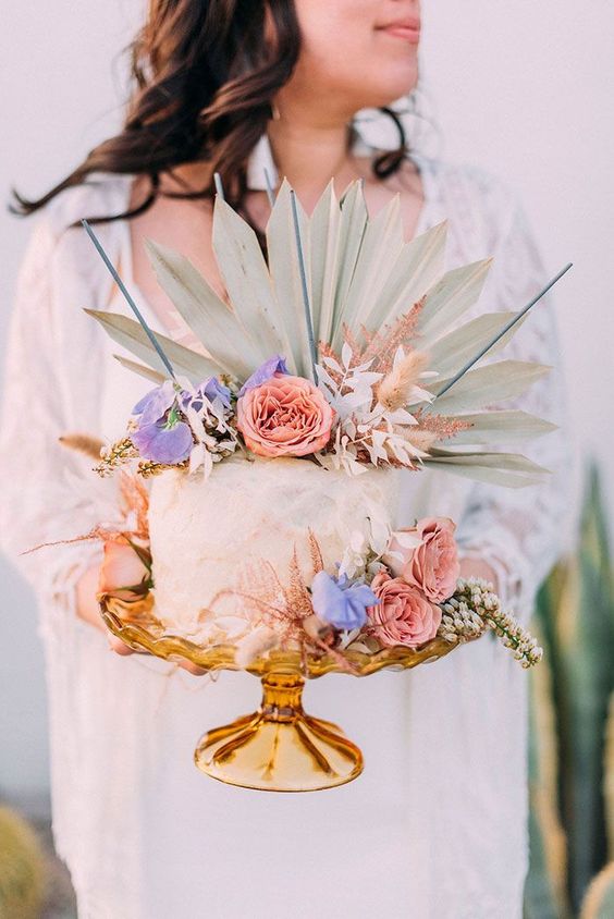a gorgeous boho wedding cake with pink and blue blooms, a frond on top and some pink leaves for a boho tropical wedding