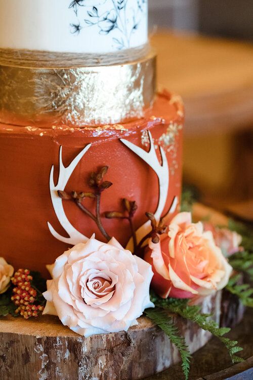 a woodland wedding cake with a white, gold leaf and rust tier, with gold sparkles, plywood antlers and fresh blooms and greenery