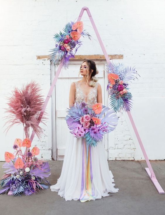 a beautiful iridescent wedding arch with a pink base, blue, purple, lilac, peahcy pink grasses and fronds is a fabulous idea