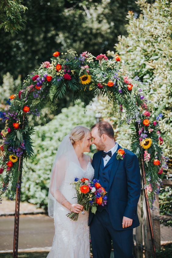 a colorful rustic wedding arch with textural greenery, bold blooms and blooming branches hanging down is a perfect fir for a colorful wedding