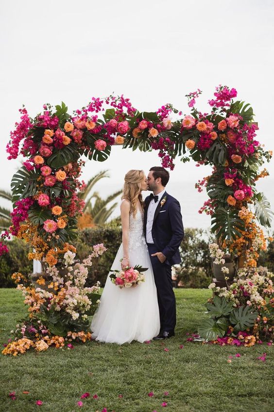 a colorful tropical wedding arch with hot pink, light pink, rust, yellow and orange blooms and monstera leaves is a gorgeous decor idea