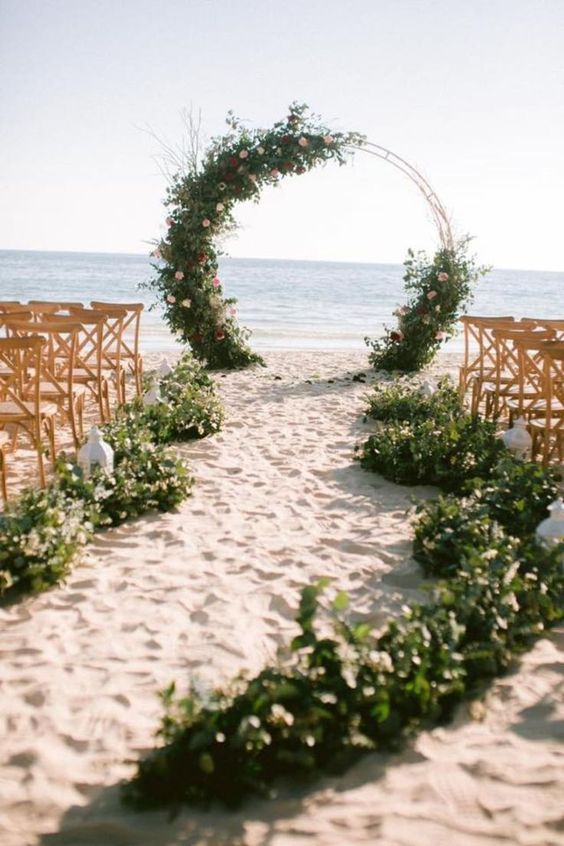 a beautiful round wedding arch covered with greenery, burgundy, blush and pink blooms and matching garlands on the sand