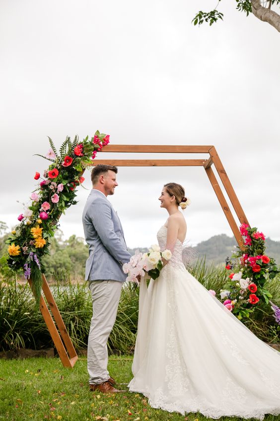 a bright hexagon wedding arch with greenery, pink, yellow and red blooms is a lovely idea for a bold tropical wedding