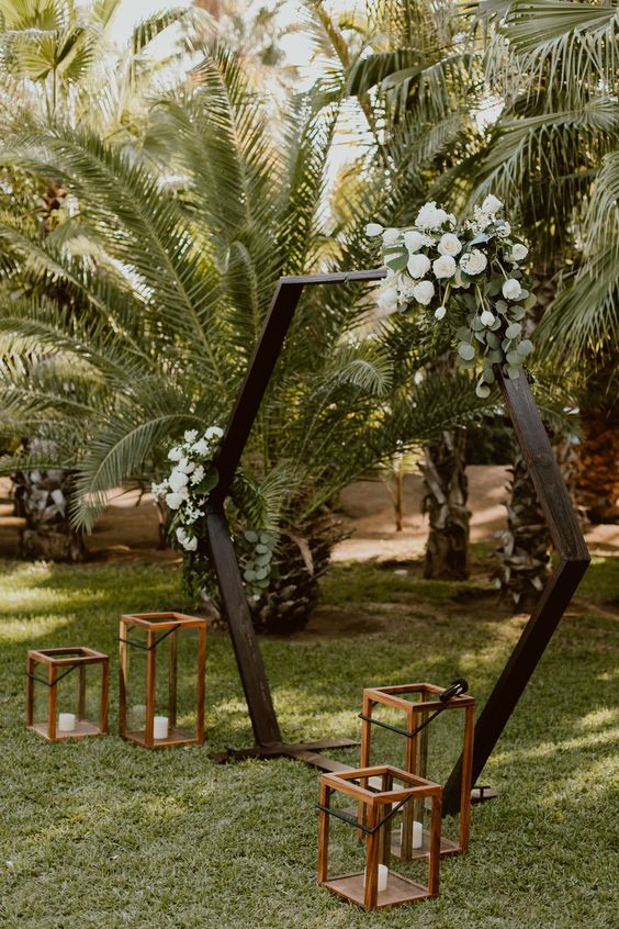 a dark stained hex wedding arch with greenery and white blooms and candle lanterns at the base for a chic wedding