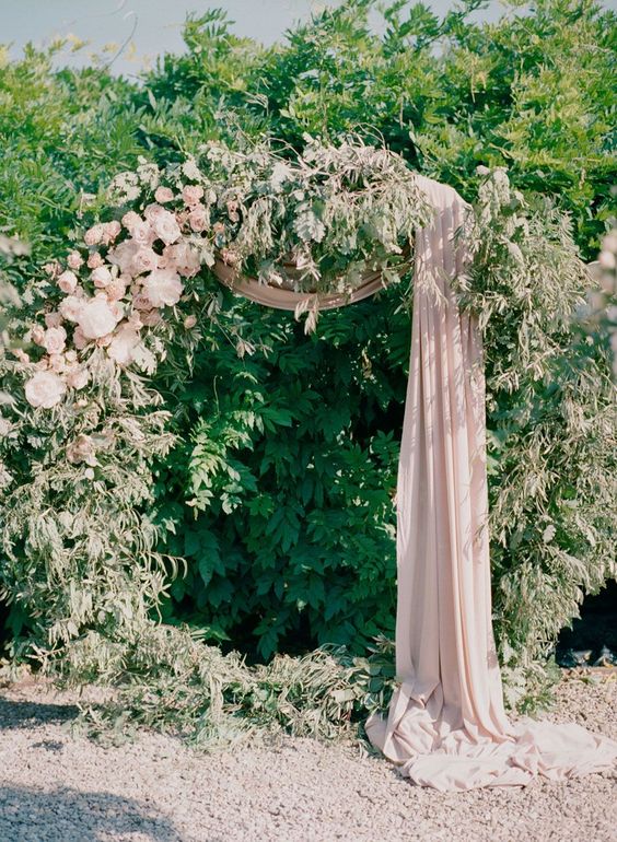 a romantic round wedding arch with greenery, blush blooms and blush fabric on the arch and the ground