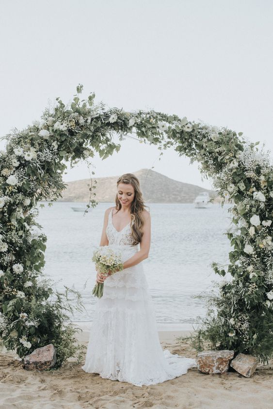 a serene round wedding arch with textural greenery, white blooms and a sea view as a backdrop