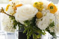 a bright summer wedding centerpiece of a clear vase, greenery, white and yellow blooms and billy balls
