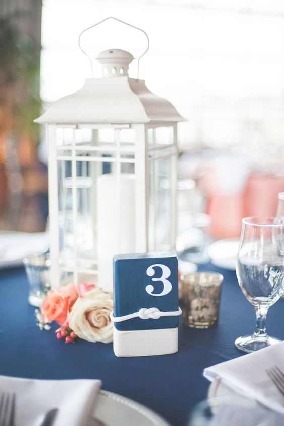 a classic nautical wedding centerpiece of a candle lantern, white and coral blooms, a table number and some candles