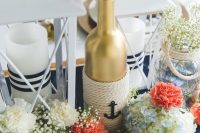 a creative nautical wedding centerpiece of a navy table runner, candle lanterns with stripes, coral and white blooms and baby’s breath