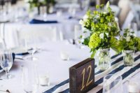 a nautical rustic wedding tablescape with a white tablecloth, a striped runner, a dark stained table number and greenery
