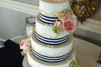 a nautical wedding cake with gold lace, navy ribbons, pink blooms and greenery and an anchor on top
