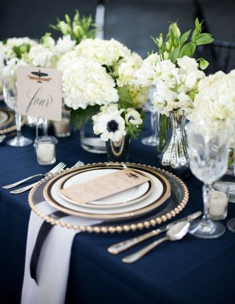 a nautical wedding table setting with a navy tablecloth, white napkins, white floral centerpieces and touches of gold