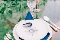 a nautical wedding tablescape with a glass table, a nautical plate, greenery and a boat, navy napkins is modern and cool