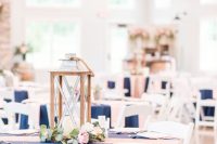 a nautical wedding tablescape with a navy tablecloth, blush napkins, a candle lantern and greenery and blooms