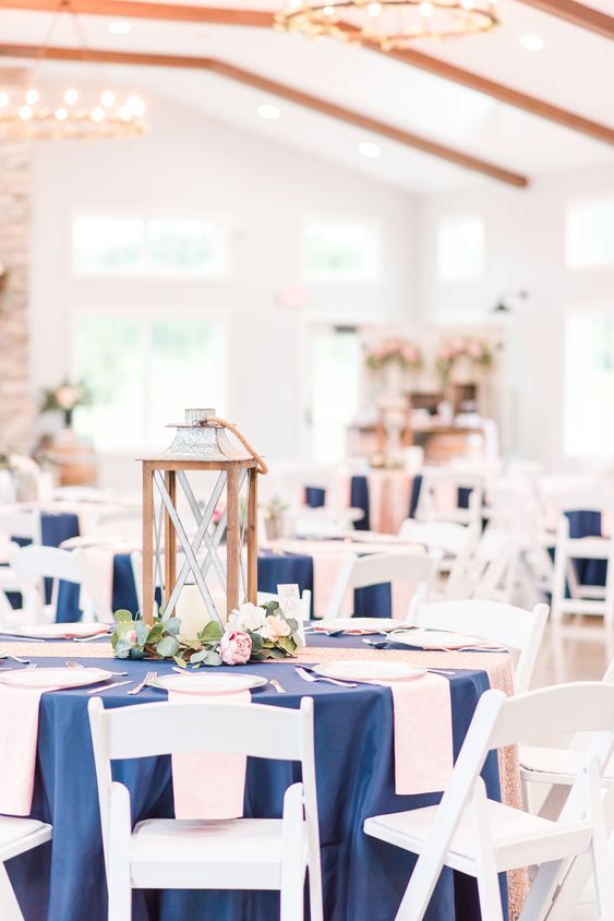 a nautical wedding tablescape with a navy tablecloth, blush napkins, a candle lantern and greenery and blooms