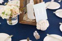a nautical wedding tablescape with a navy tablecloth, white plates and silver, a wooden candle lantern, neutral blooms and a roper knot