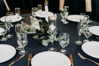 a refined and elegant nautical wedding tablescape with a navy tablecloth, white napkins, greenery and a cube plus candles