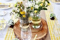 a rustic wedding centerpiece of jars with greenery, white and yellow roses and billy balls on a wood slice is cool for spring