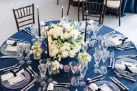 a sophisticated nautical wedding tablescape with a navy tablecloth, white napkins and menues, navy candles and a lush floral centerpiece
