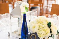 a stylish nautical wedding centerpiece of a candle lantern, a navy bottle with white blooms and a matching centerpiece in a vase, a navy wood and rope table number