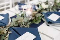 an elegant nautical wedding table setting with a navy runner and napkins, a greenery runner and candles, a silver charger and cutlery