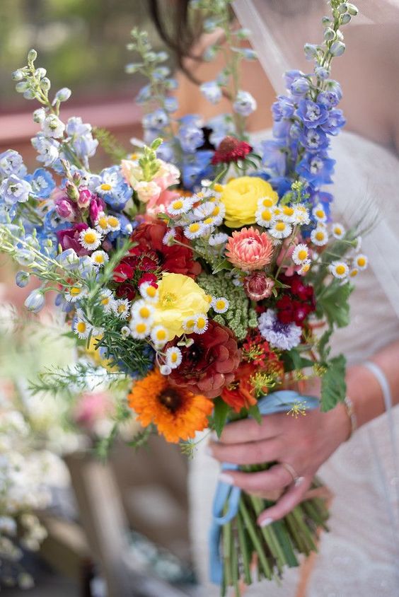 a colorful summer wedding bouquet that includes orange, yellow, burgundy, blue and purple blooms and some daisies as fillers