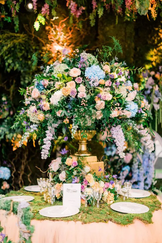 a beautiful and lush wedding centerpiece of pink, peachy, blue blooms, lots of greenery and a gilded vase is an amazing secret garden wedding centerpiece