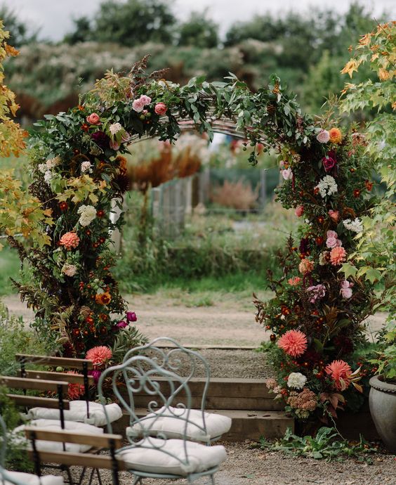 a beautiful secret garden wedding arch covered with leaves, blush, white, deep and bright red blooms is a very chic and bold idea for fall