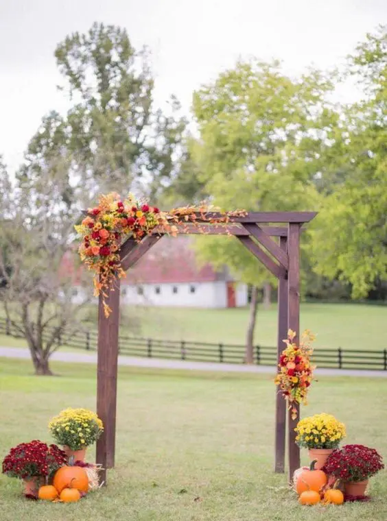 a bold rustic wedding arch of dark stained wooden slabs, bright blooms and greenery and pumpkins and flowers around