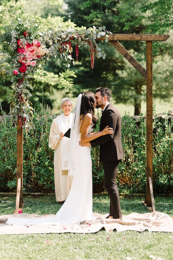 a bold rustic wedding arch of rich stained wood, greenery, pink, hot pink blooms and a rug on the ground is amazing