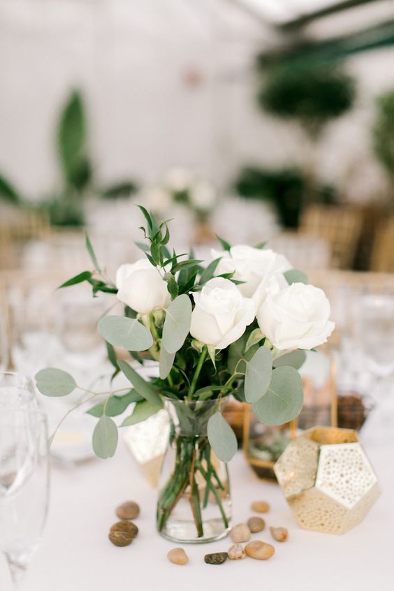a chic and simple wedding centerpiece of a vase, white roses and greenery, pebbles and a faceted candleholder