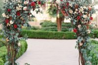 a chic fall wedding arch covered with greenery, blush and red blooms plus some twigs for a more eye-catchy looks is a very cool idea for an autumen garden wedding