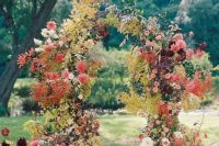 a colorful fall garden wedding arch decorated with greenery and yellow fall leaves, red, burgundy, peachy pink blooms and white flowers