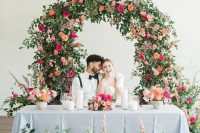 a colorful garden wedding arch covered with greenery and super bold blooms and matching floral arrangements at the table